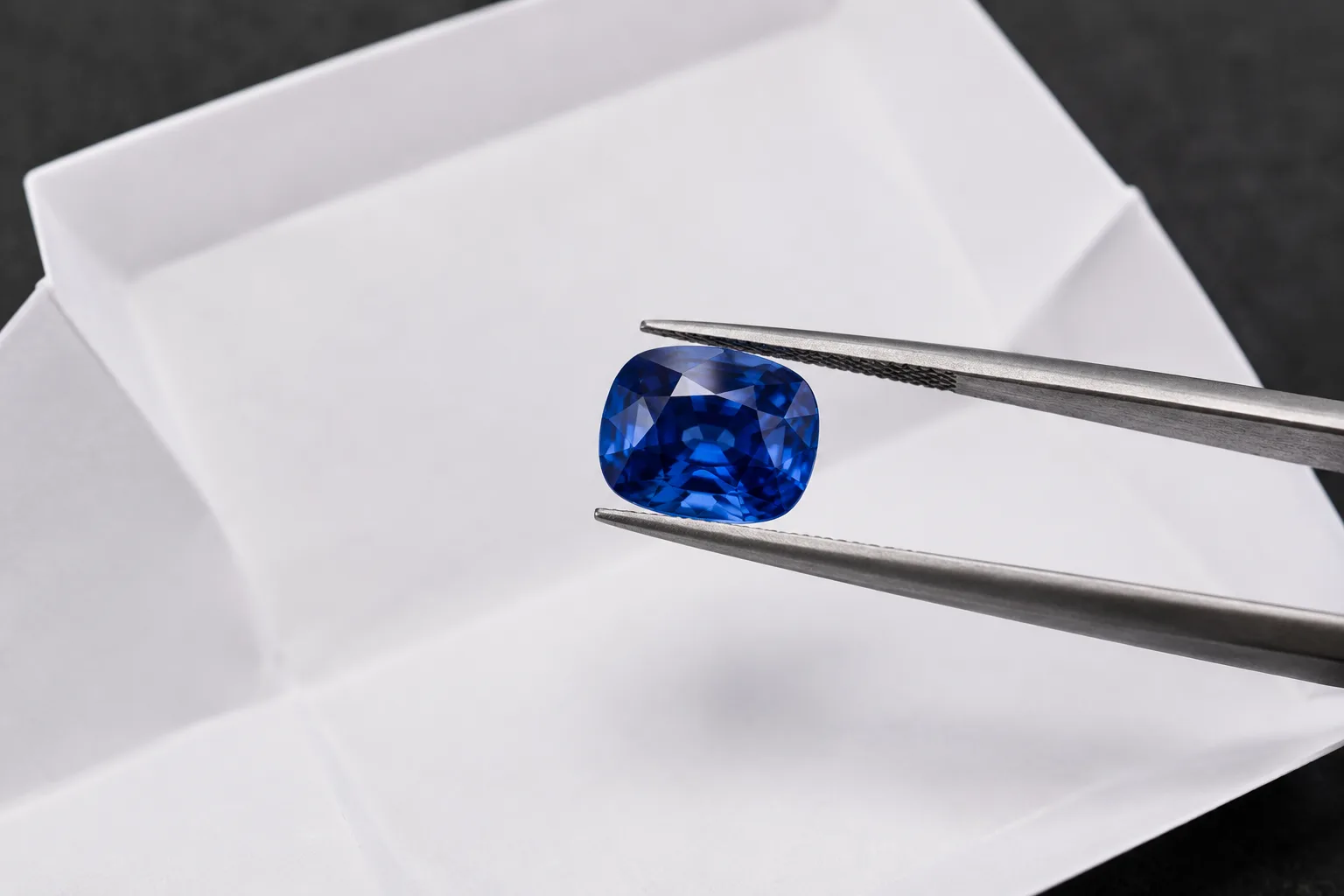 A faceted blue sapphire held in steel tweezers over folded white gem paper