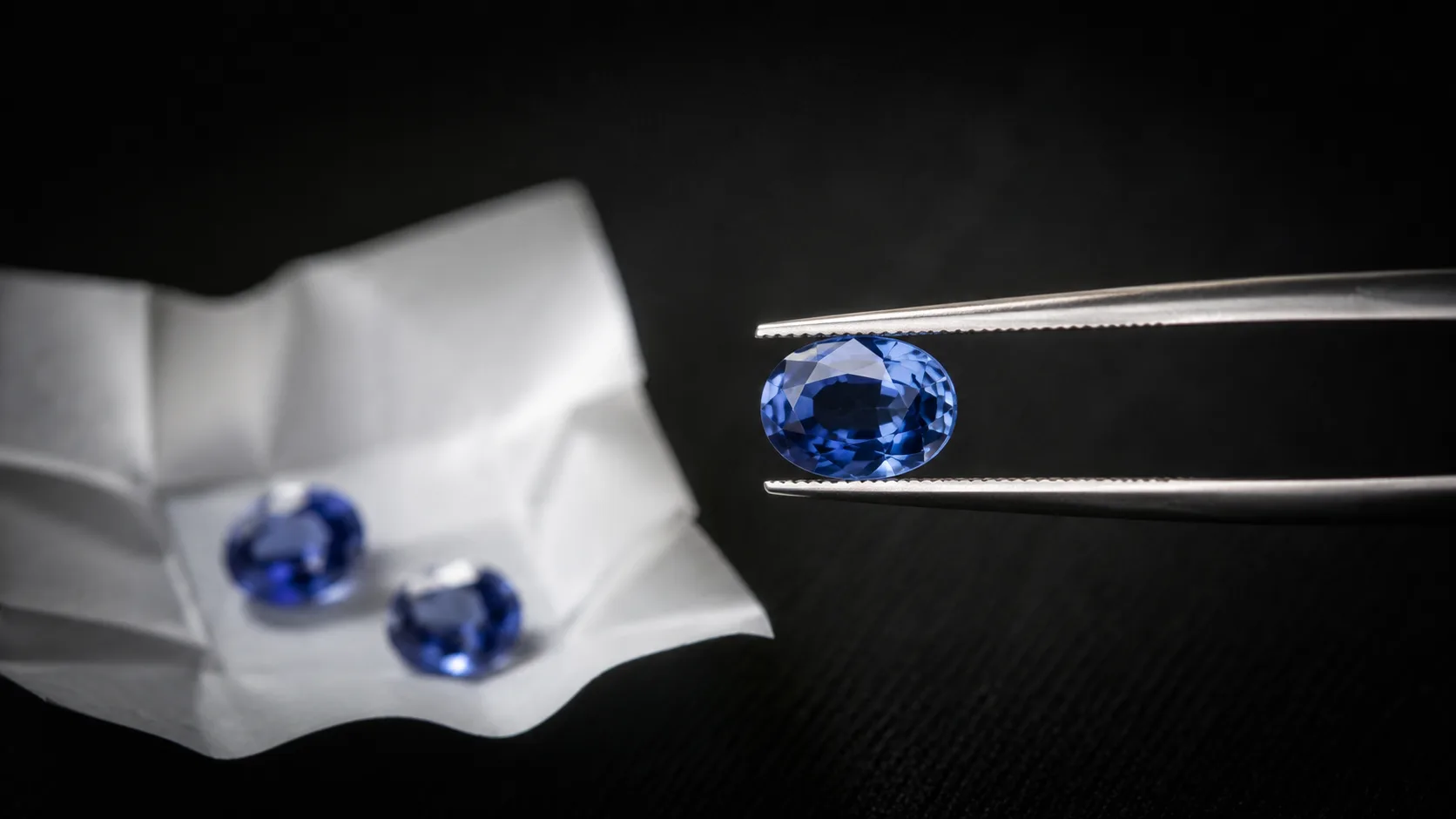 Oval blue Ceylon sapphire held in steel tweezers beside folded white gem paper with two loose sapphires blurred behind