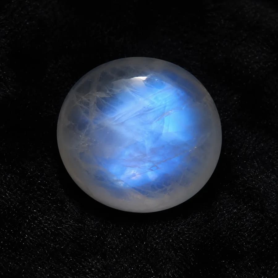 Round moonstone cabochon with soft blue adularescence glowing across the dome, on black cloth