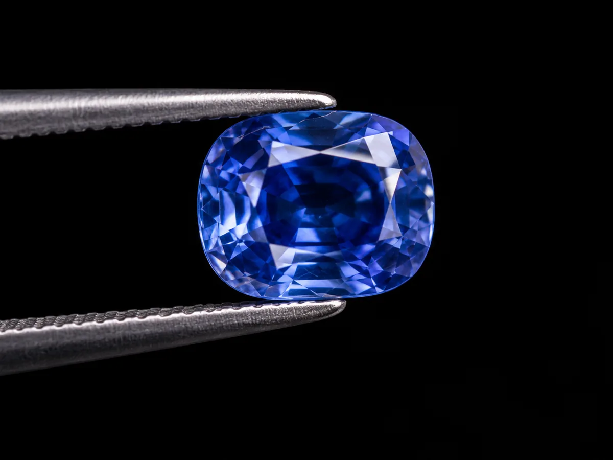 Tray of faceted coloured gemstones with a blue sapphire held in tweezers
